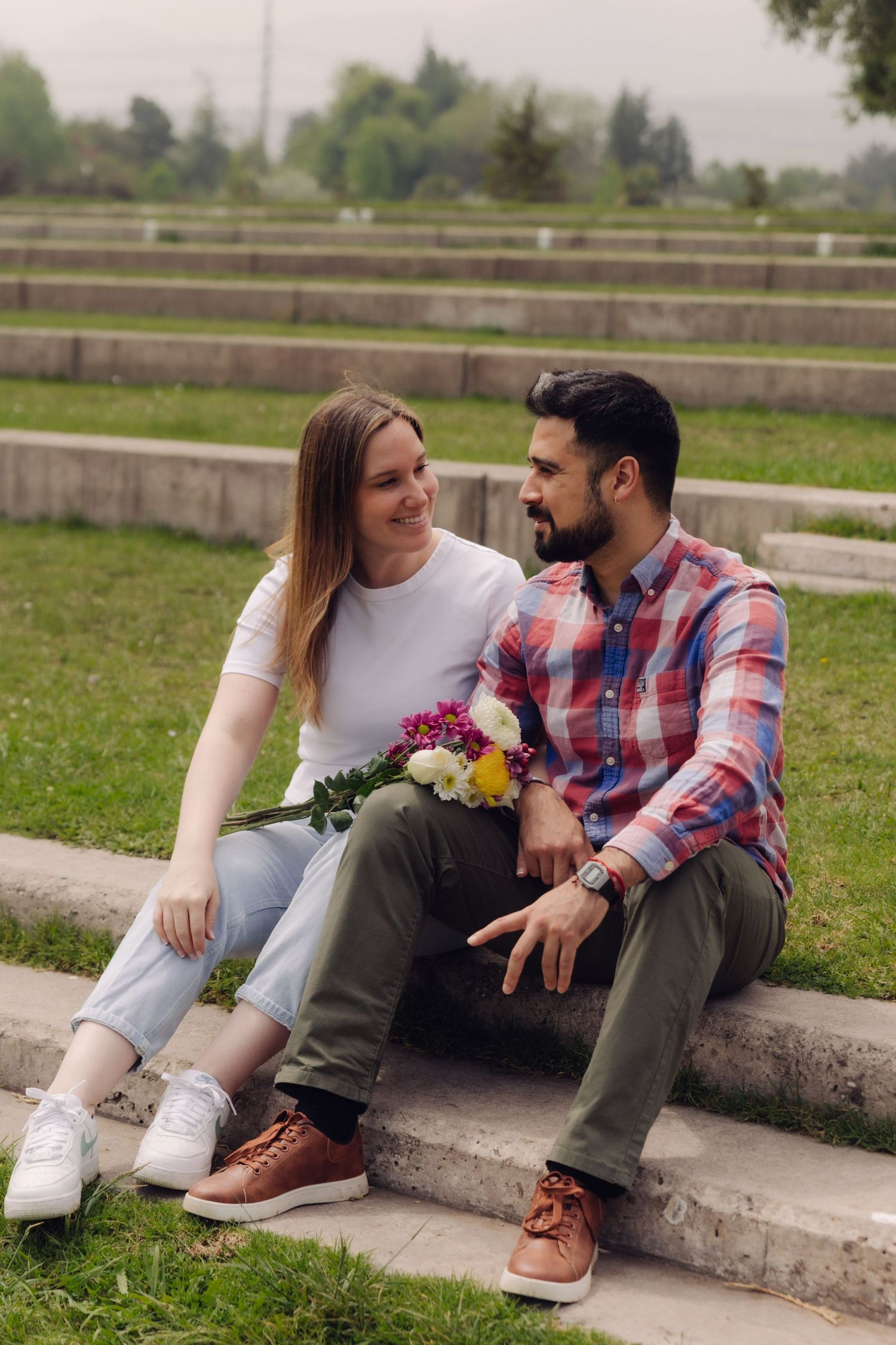 Couple sitting on stone steps in a park, sharing a bouquet of colorful flowers together.