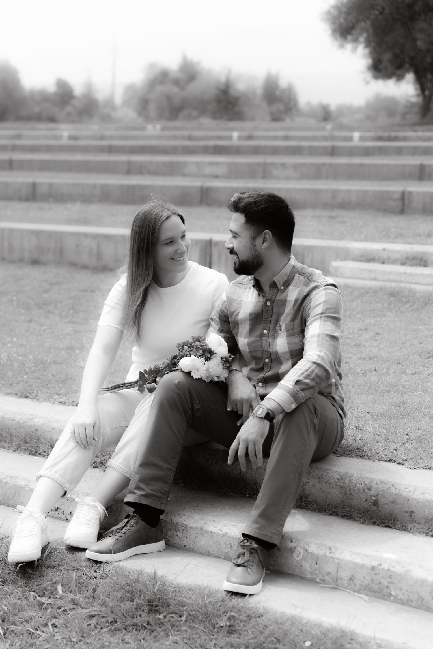 Black and white photo of a couple sitting on steps in a park, smiling at each other.
