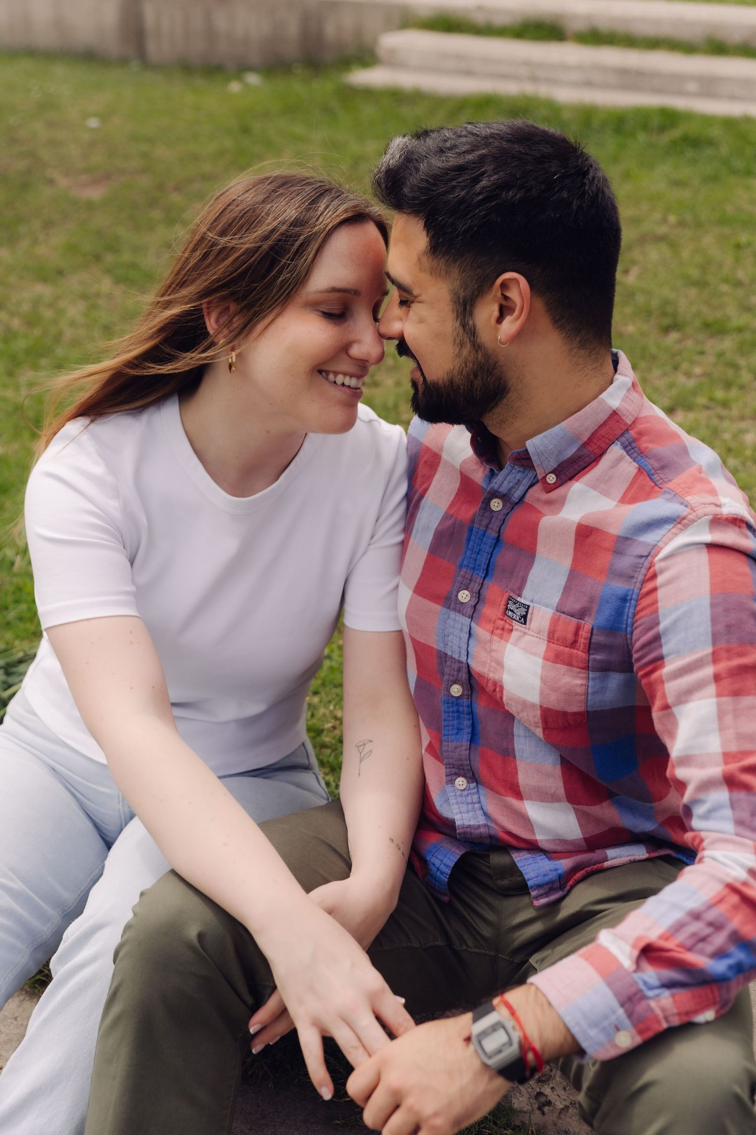 Smiling couple touching foreheads romantically while sitting on grass in a park, woman in white tee and man in plaid shirt.