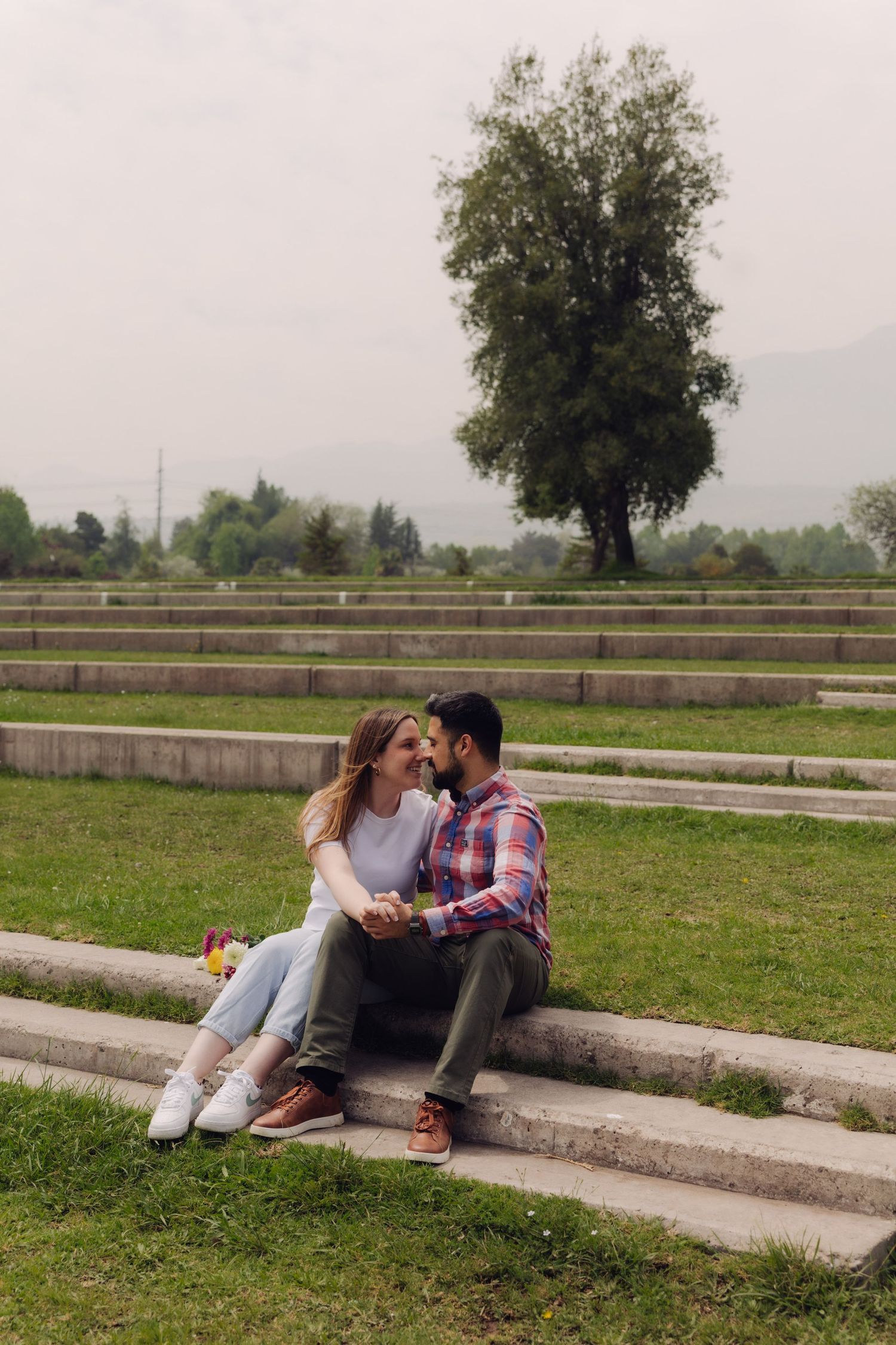 Happy couple sitting on outdoor stone steps holding hands, with a large tree and misty mountains in the background.