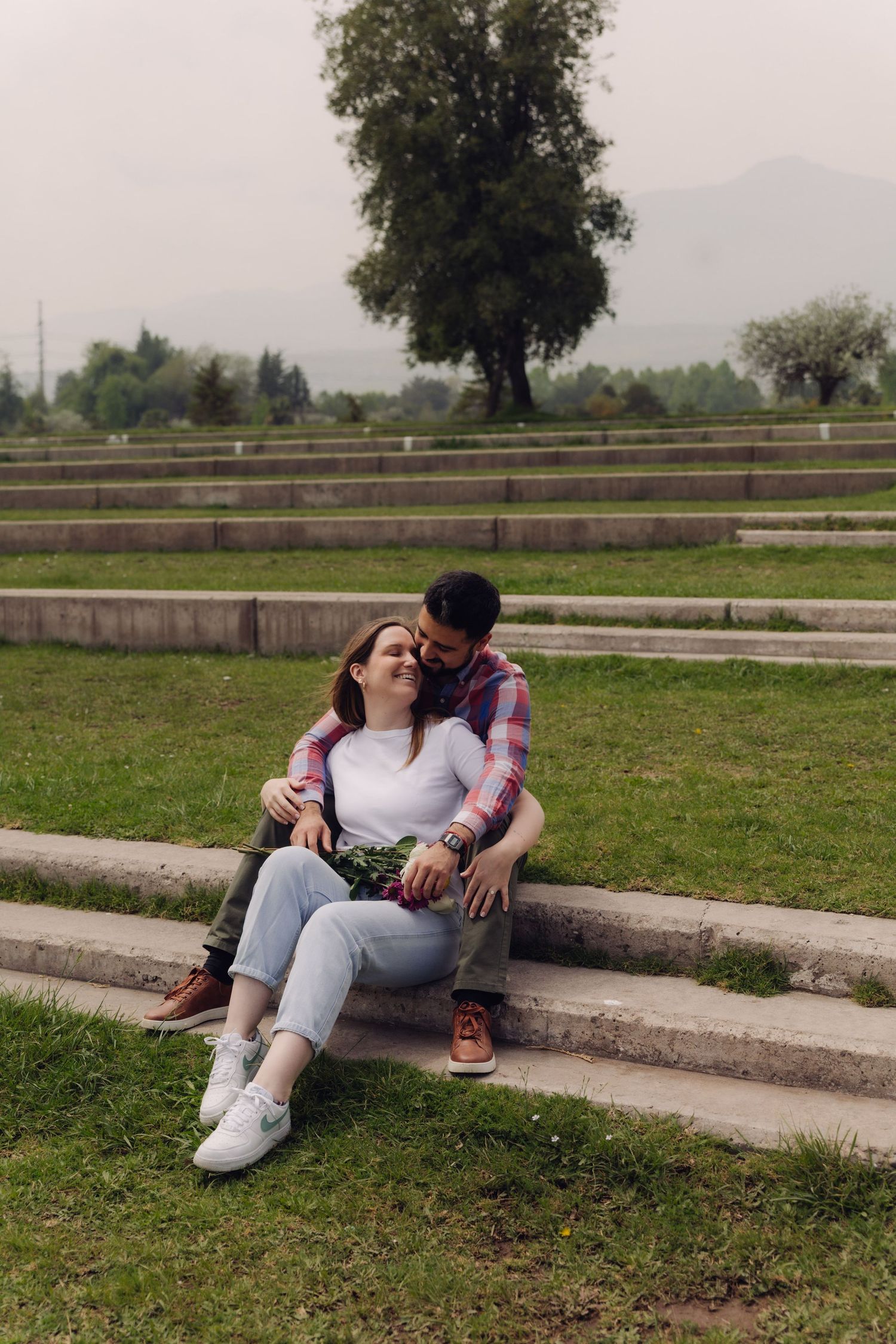 Couple sitting on stone steps in a park, man embracing woman from behind while she laughs, holding a small bouquet.