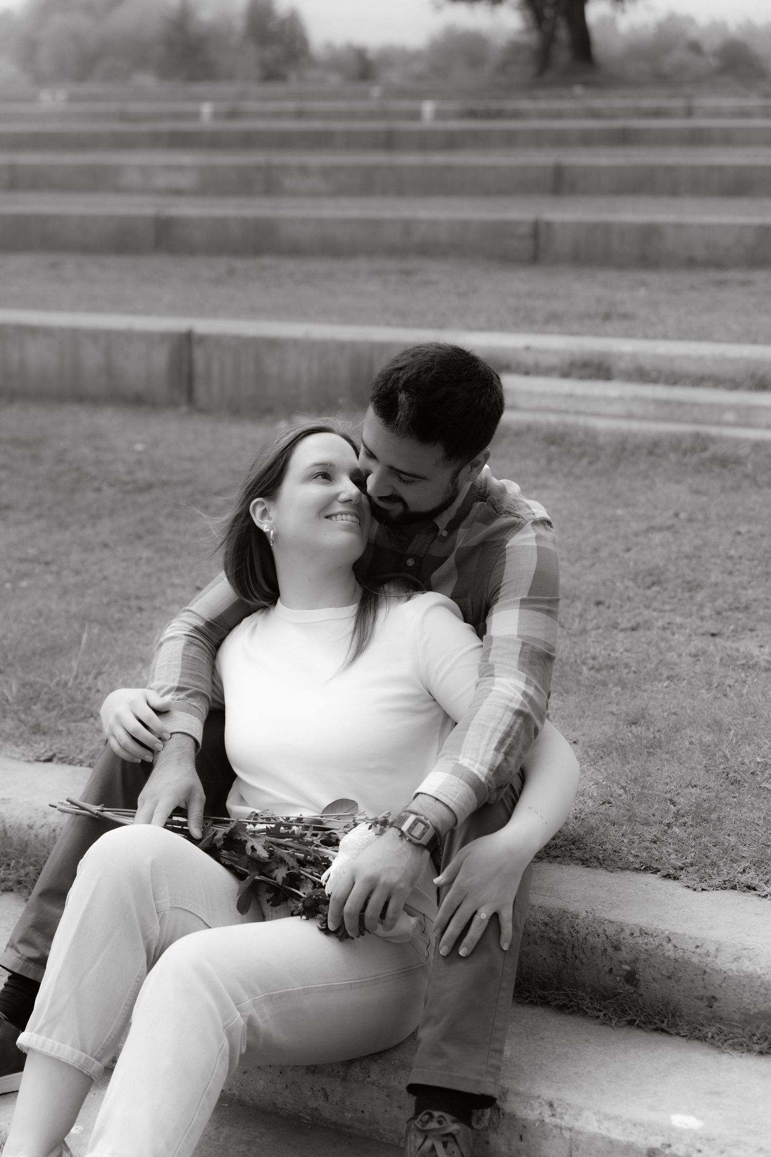 Black and white photo of a couple embracing on stone steps in a park, woman smiling up at man affectionately.