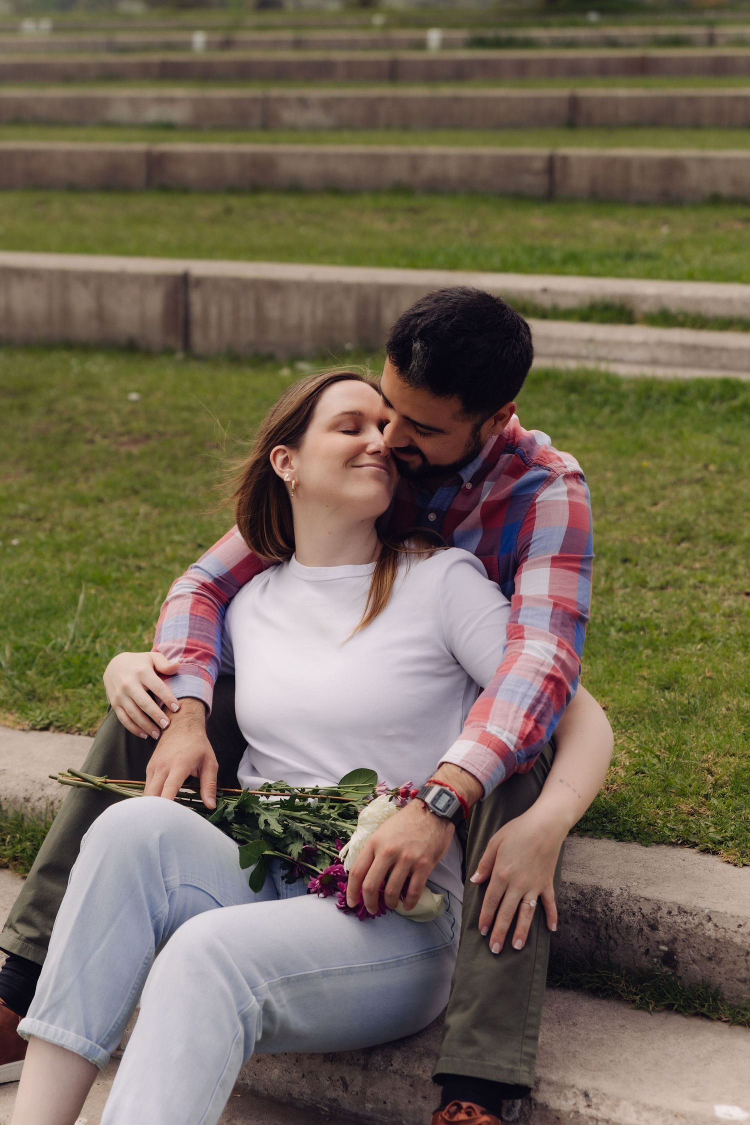 Man tenderly kissing woman on cheek while embracing her on stone steps, woman holding a bouquet of pink flowers.