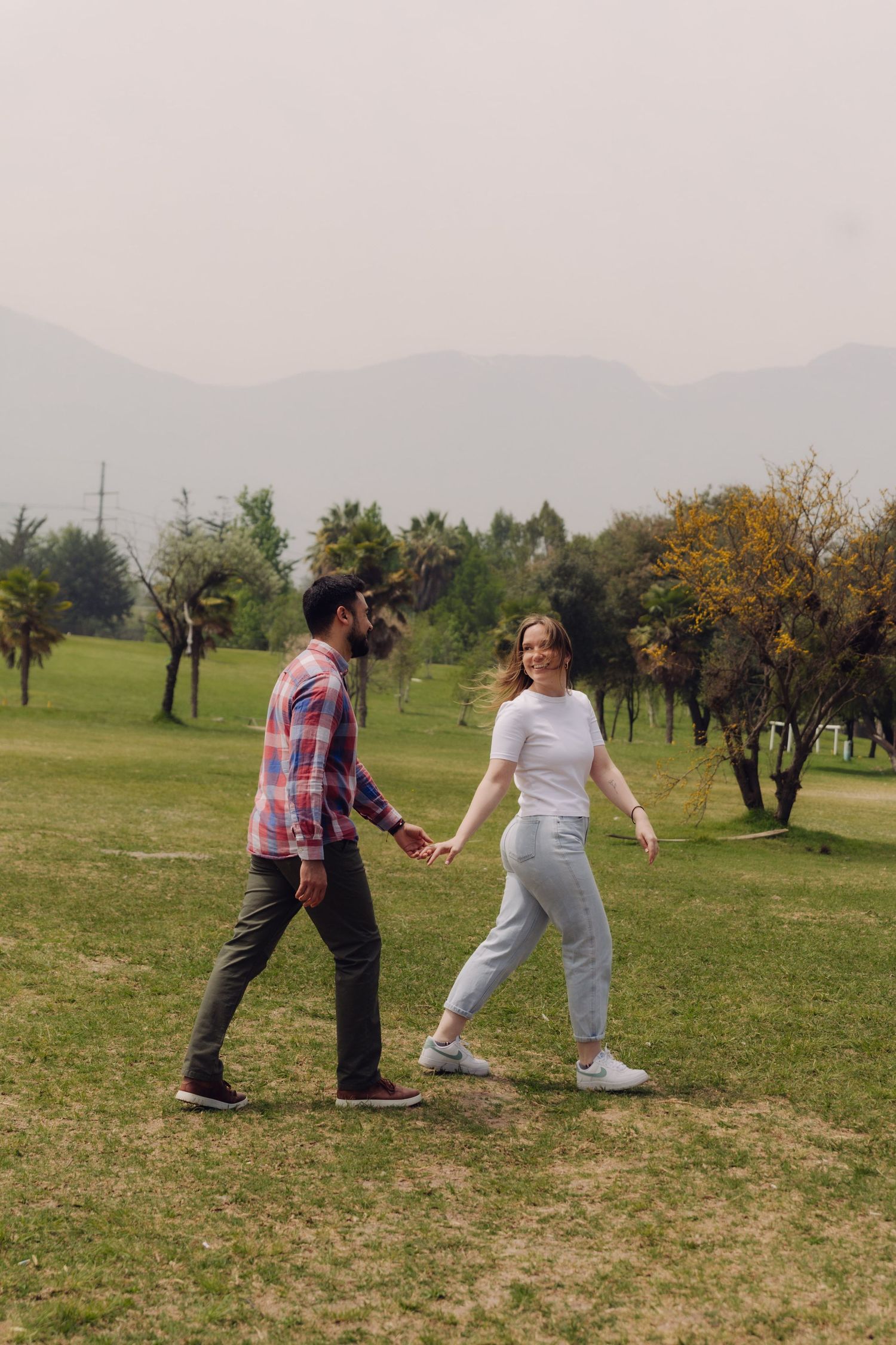Couple walking hand in hand across a green park lawn with mountains and trees visible in the misty background.