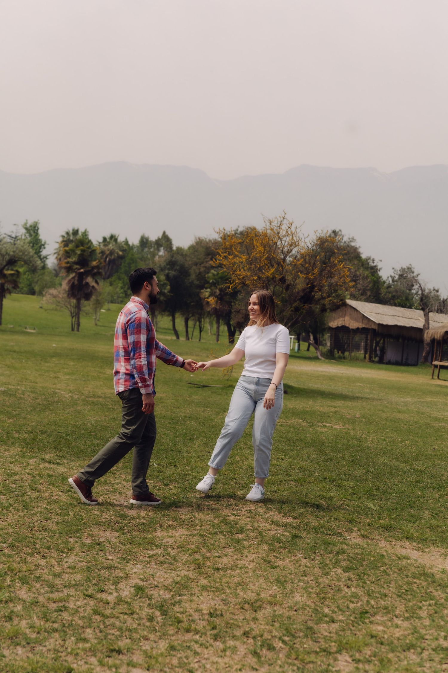 Smiling couple holding hands facing each other on a green lawn with trees, mountains, and a rustic building behind them.