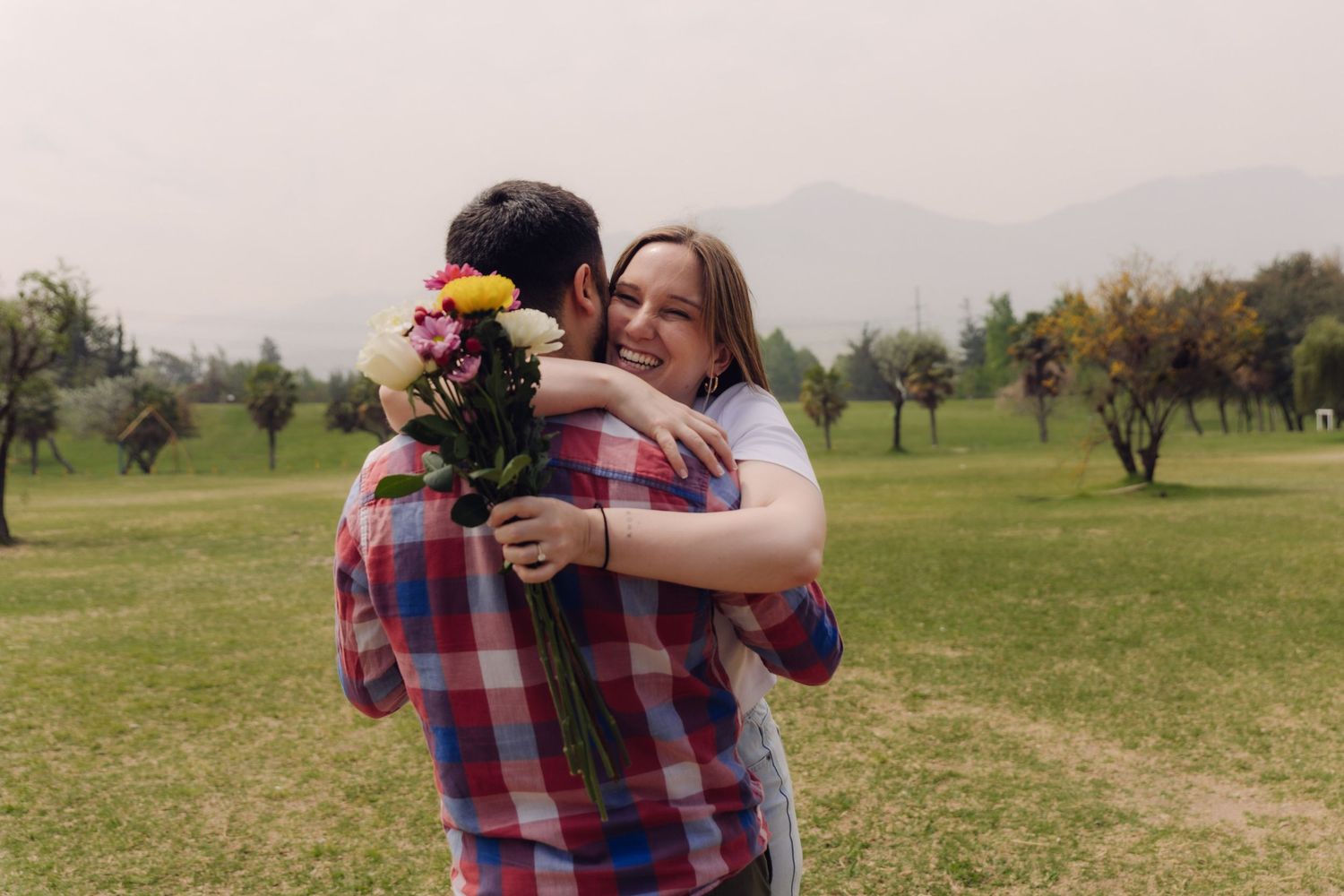 Woman joyfully hugging a man who is holding a colorful bouquet of flowers in a green park with mountains behind.
