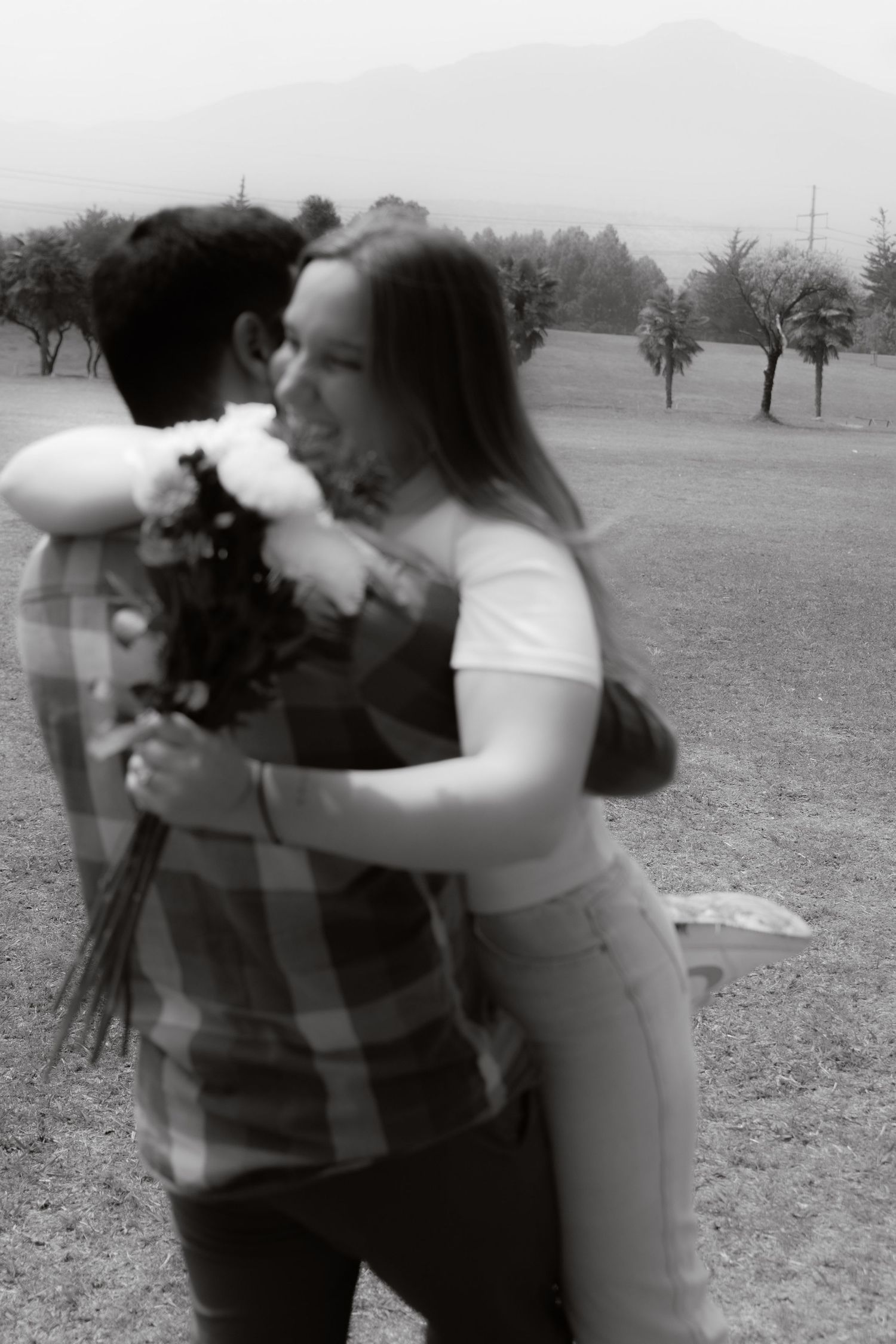 Blurred black and white artistic photo of couple embracing with flowers in an open field with mountains in background.