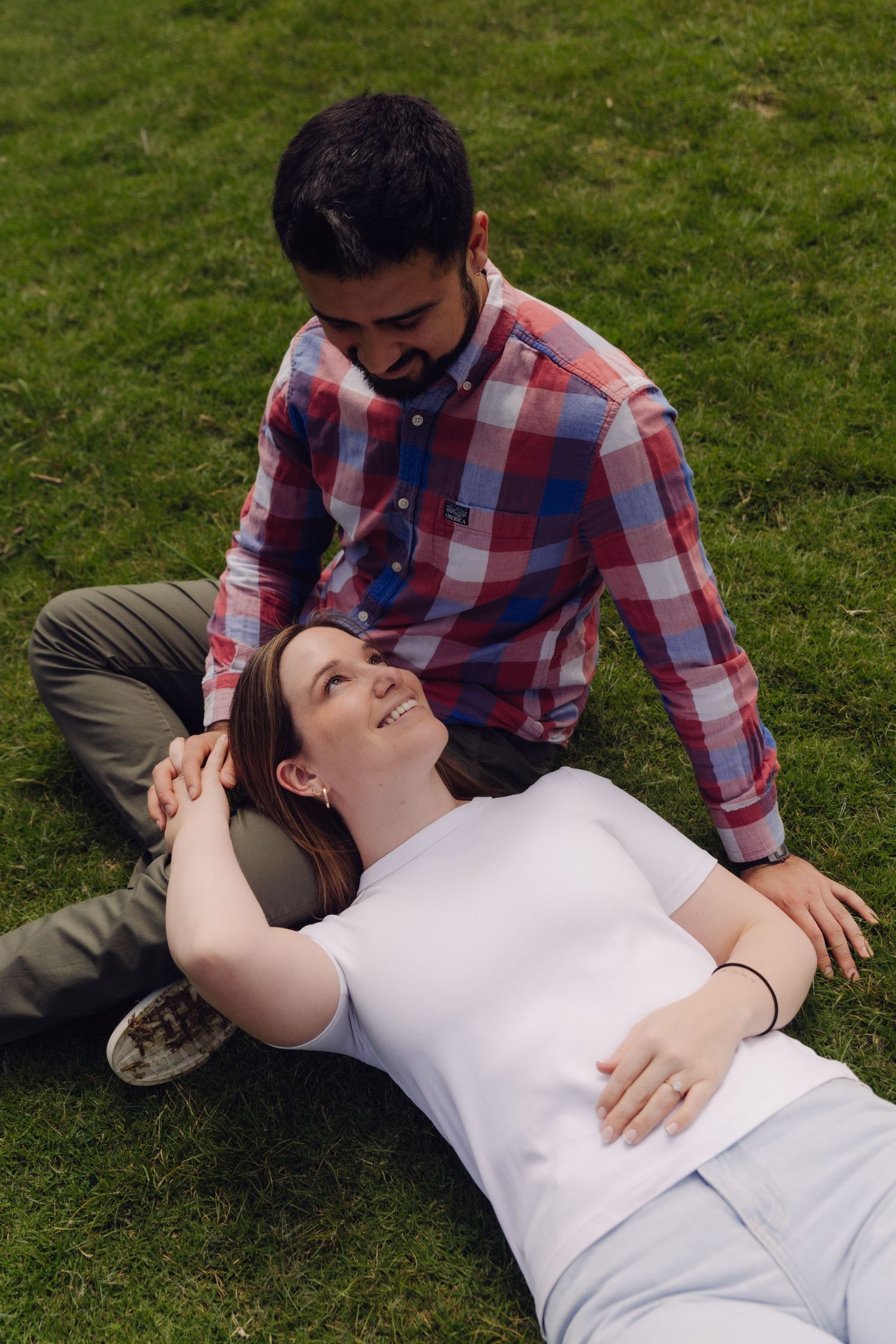 A couple relaxing on green grass, woman lying down smiling up at bearded man in plaid shirt.