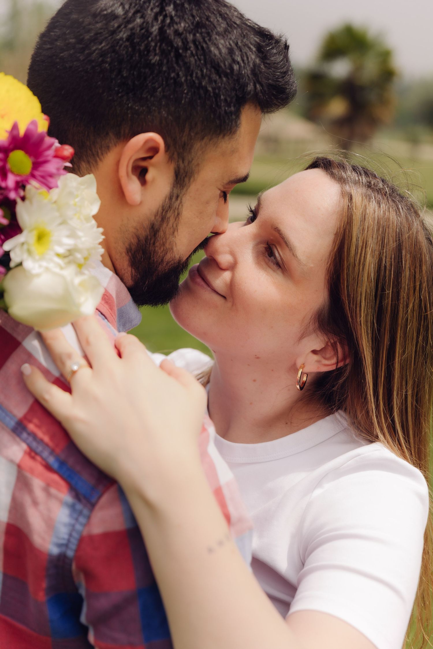 Close-up of smiling couple about to kiss, woman holding vibrant bouquet of flowers outdoors.