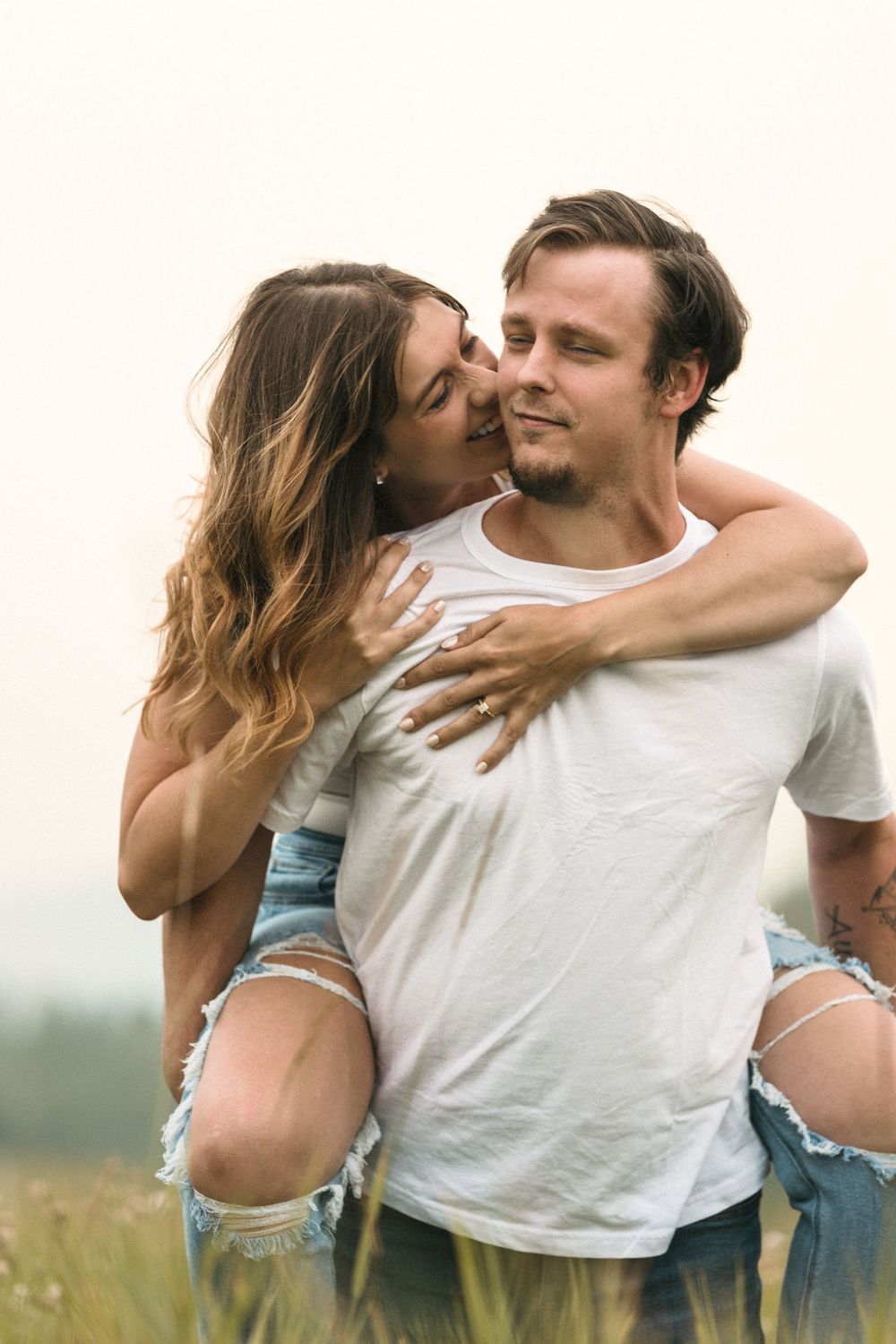 A couple shares a romantic piggyback ride in a grassy field, woman kissing man's cheek outdoors.