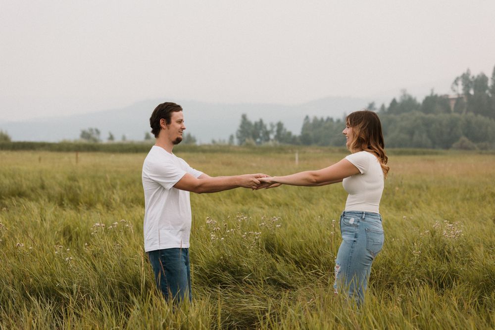 A young couple in white shirts and jeans holding hands in a lush green meadow with mountains in the background.