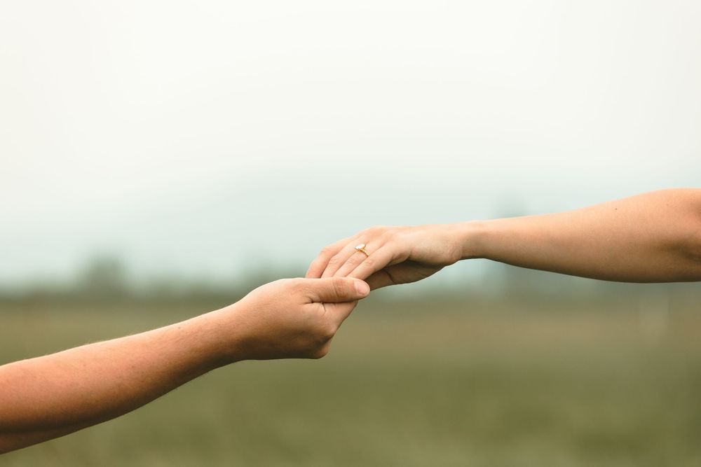 Two people holding hands outdoors with a diamond engagement ring visible on one hand.