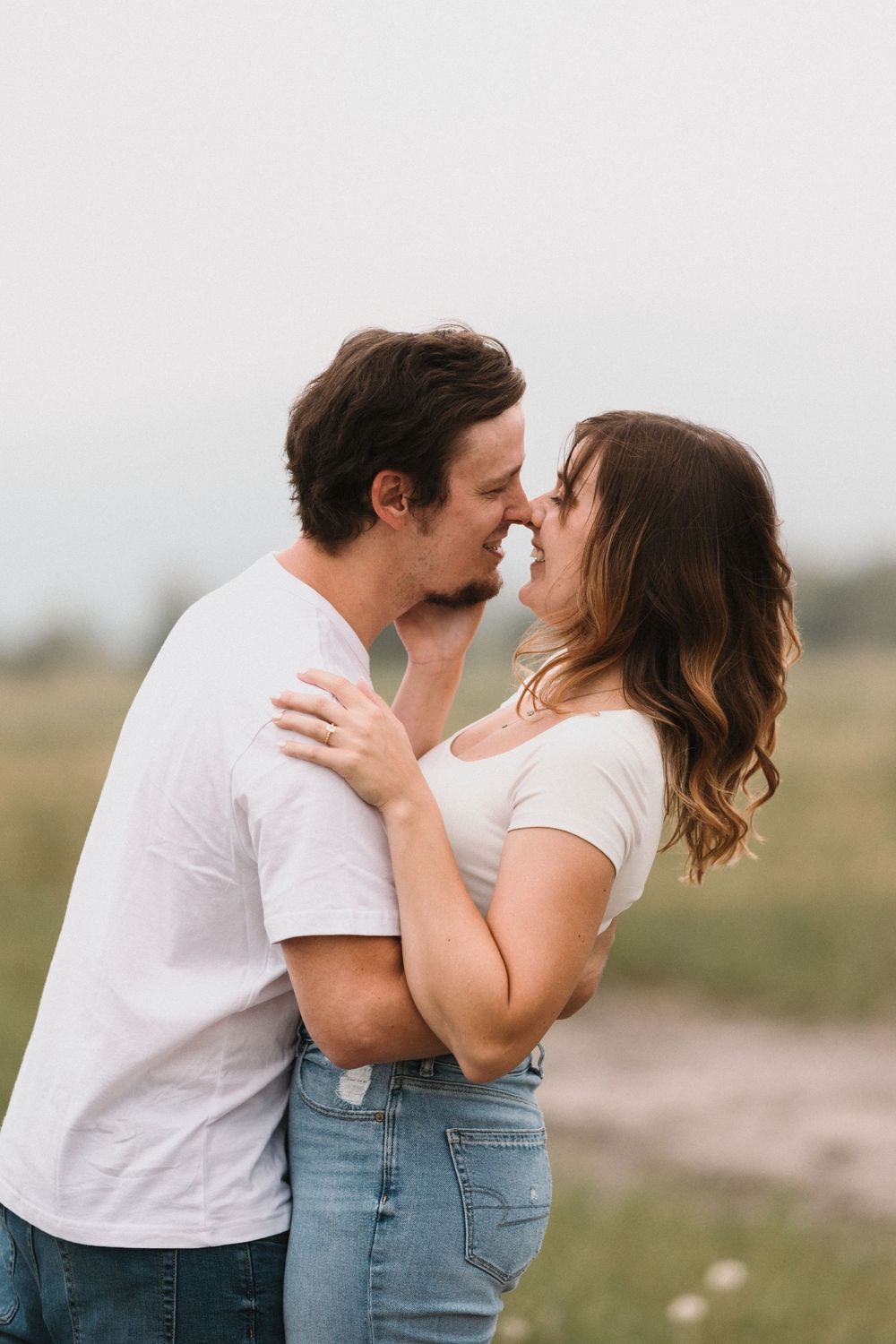 A couple in white shirts and jeans share a romantic kiss in an outdoor field setting.