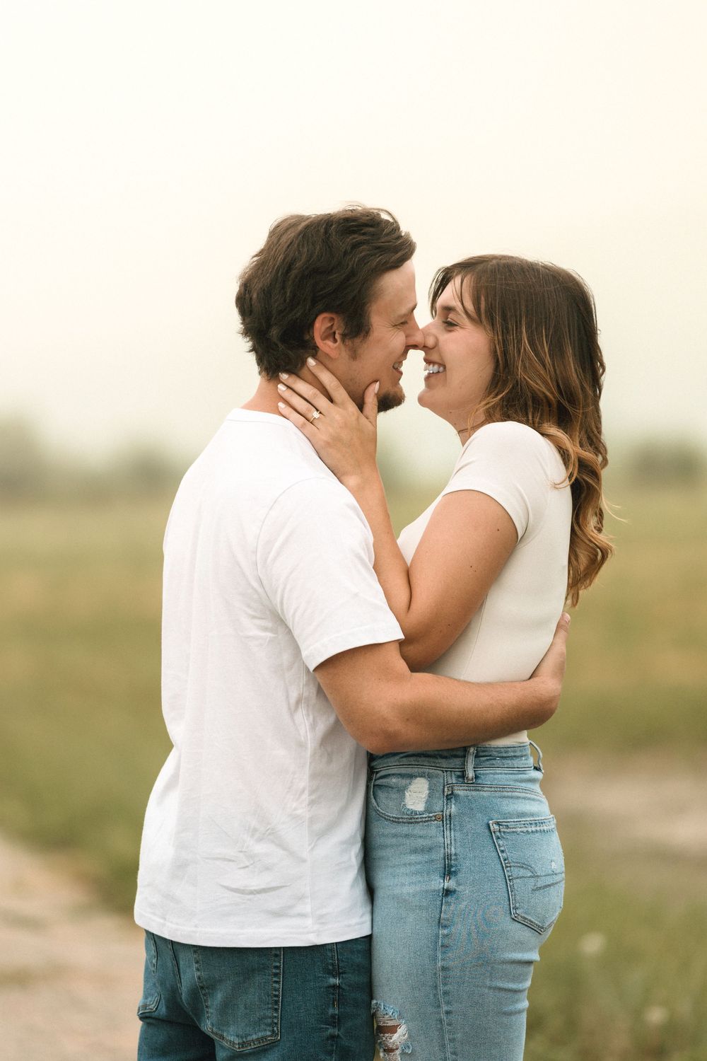 A couple in white shirts and jeans share a romantic moment outdoors, leaning in for a kiss in a scenic field.