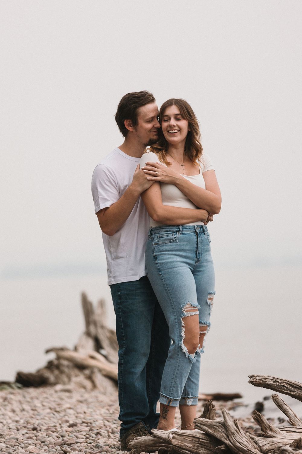 Couple embracing on a rocky beach, man kissing woman's cheek, both wearing white tops and blue jeans, overcast sky.