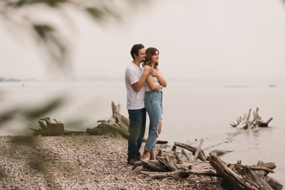 Couple embracing on a rocky shoreline with driftwood, sharing a tender moment by the calm water.