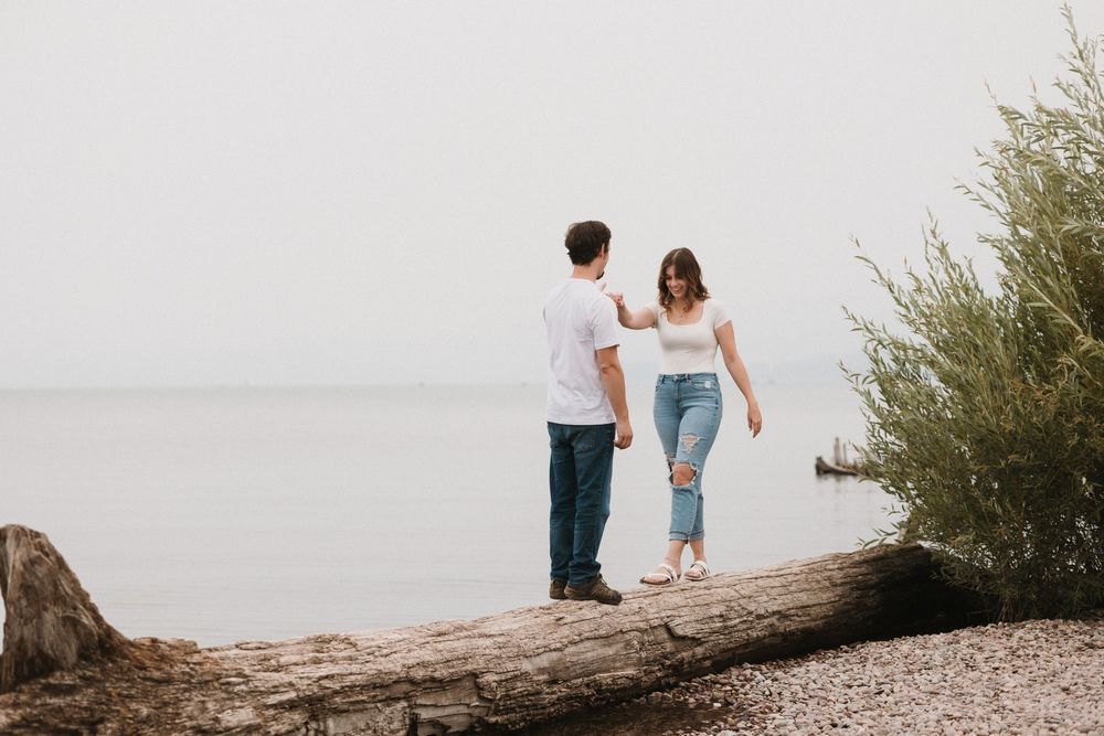 Couple balancing on a fallen log by a calm lakeside shore surrounded by lush greenery.