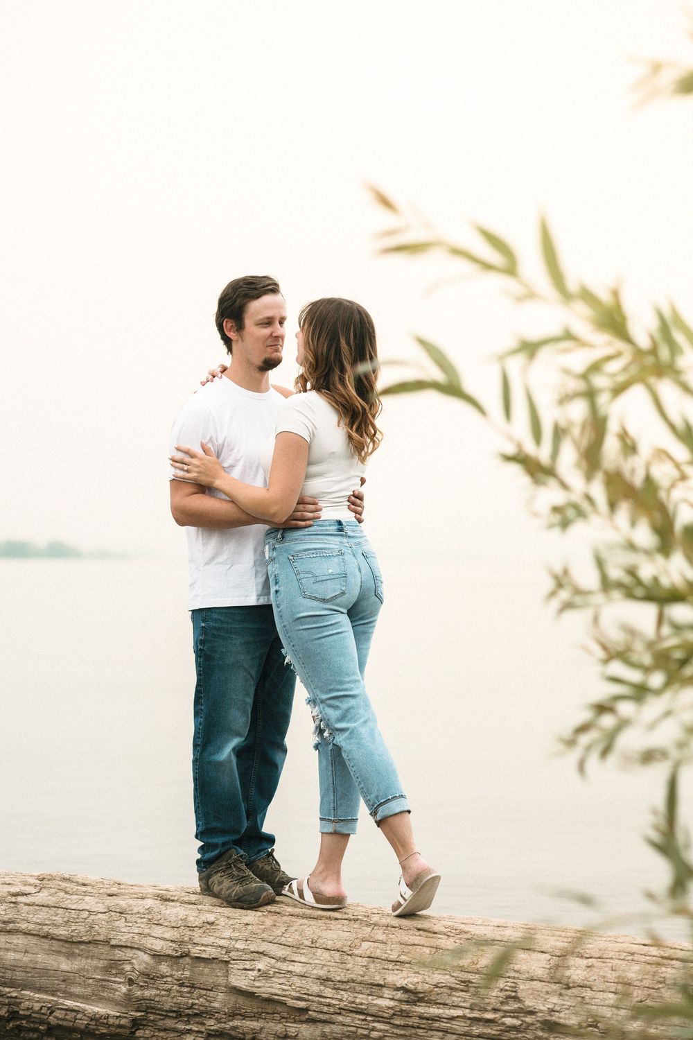 Couple in white shirts and jeans embracing on a log by a serene lake, sharing a romantic outdoor moment.