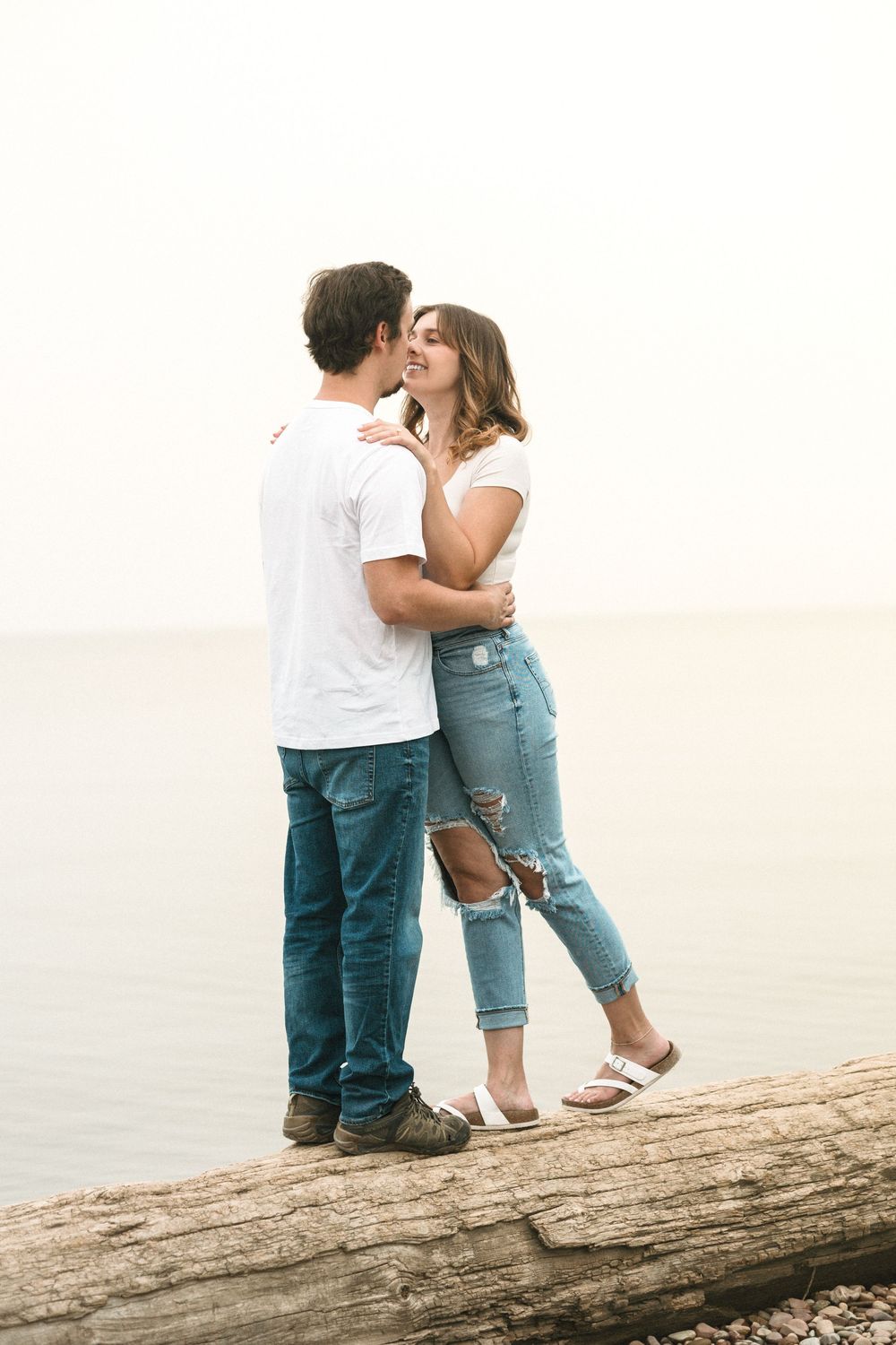 Couple in white t-shirts and jeans sharing a romantic moment on a log by the water's edge.