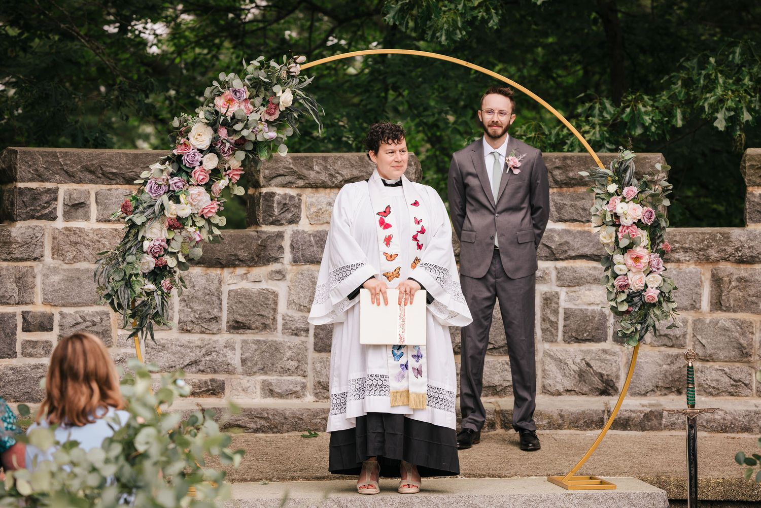Groom standing at the ceremony arch with officiant Joy Caires, whose stole is embroidered with colorful butterflies.