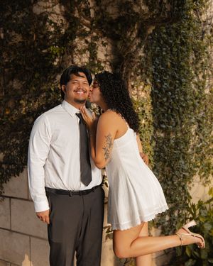 Couple sharing a romantic kiss outdoors, man in white shirt and tie, woman in white mini dress.