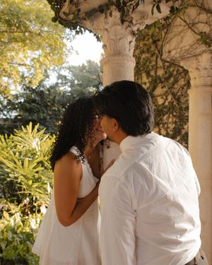 Couple sharing a romantic kiss near an ornate white column surrounded by lush greenery.