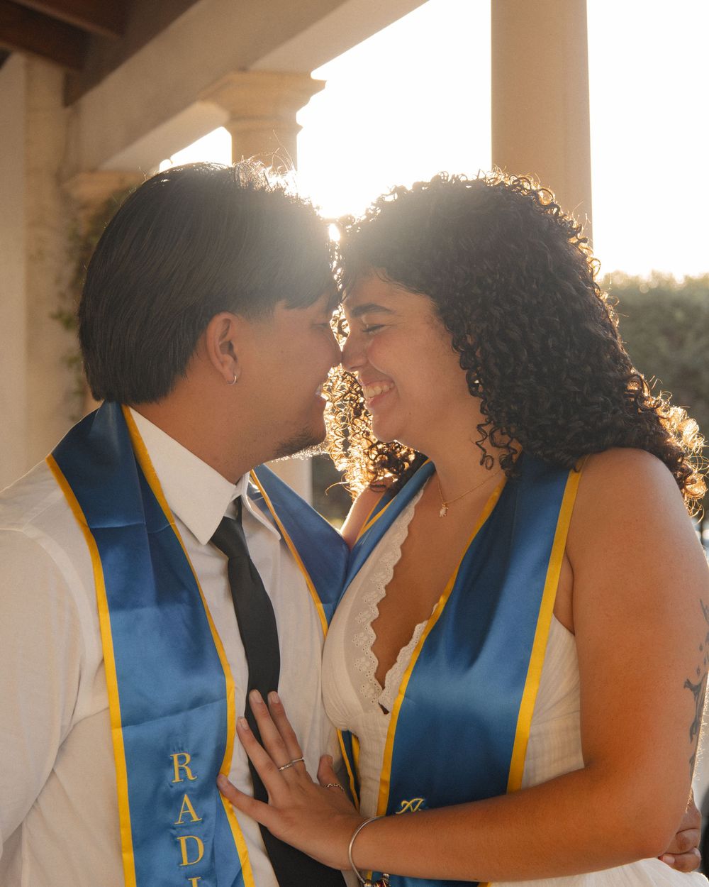 Two graduates in blue and gold stoles sharing a joyful moment at golden hour.