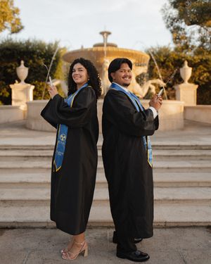 Couple in black graduation gowns and blue stoles posing back-to-back near a fountain.