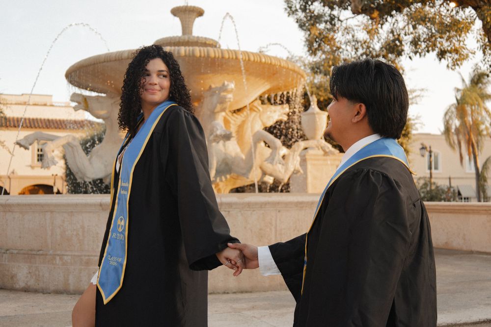 Couple in graduation gowns holding hands in front of an ornate stone fountain.
