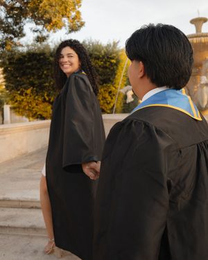 Graduate in black gown smiling back at partner near a decorative outdoor fountain.