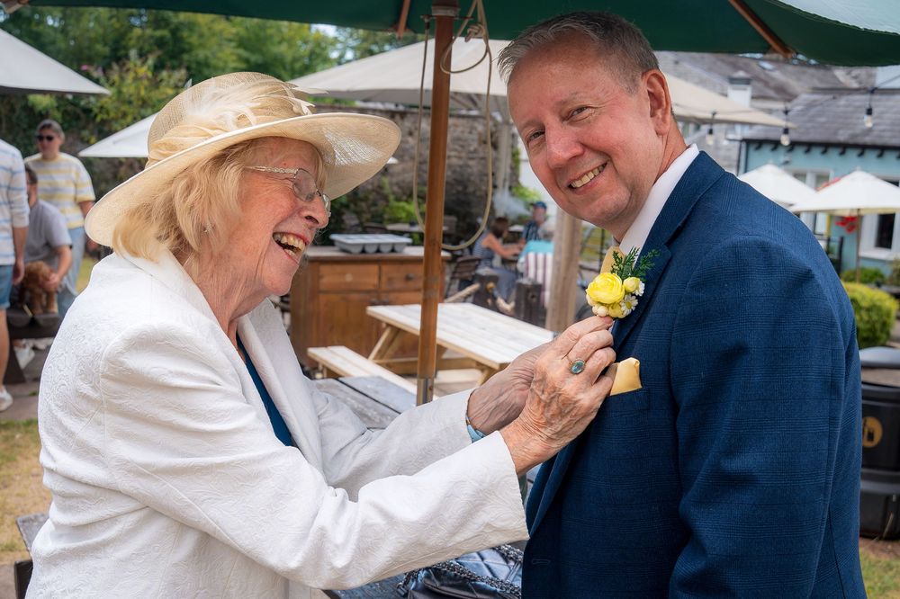 Elderly woman in sun hat pins boutonniere on smiling man in blue suit at outdoor wedding celebration.