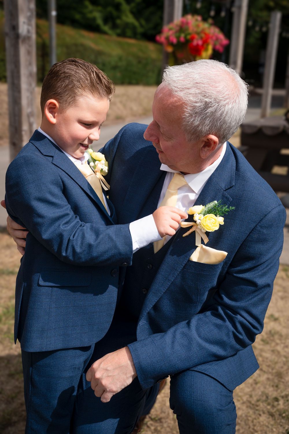 A boy and older man in matching blue suits with yellow boutonnieres share a tender moment at a wedding.