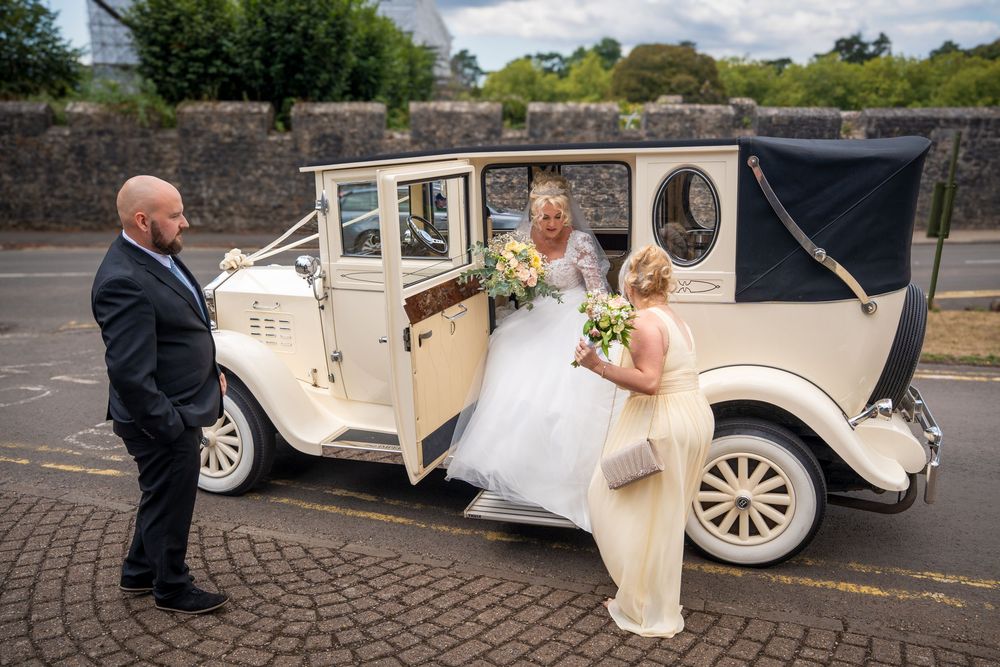 Bride stepping out of vintage ivory wedding car, greeted by groom and bridesmaid holding bouquets outdoors.
