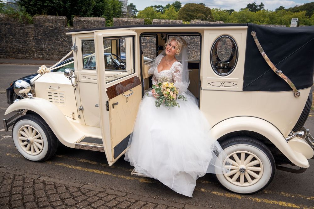 Bride in white gown posing beside a vintage cream wedding car, holding a bouquet on her special day.