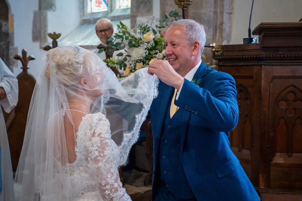 Groom in blue suit lifting bride's veil at church wedding ceremony altar.