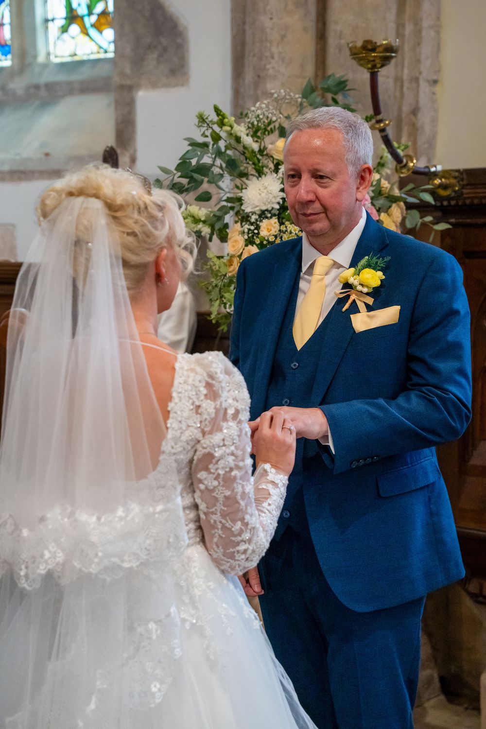 Groom in blue suit placing wedding ring on bride's finger during church ceremony with floral arrangements.
