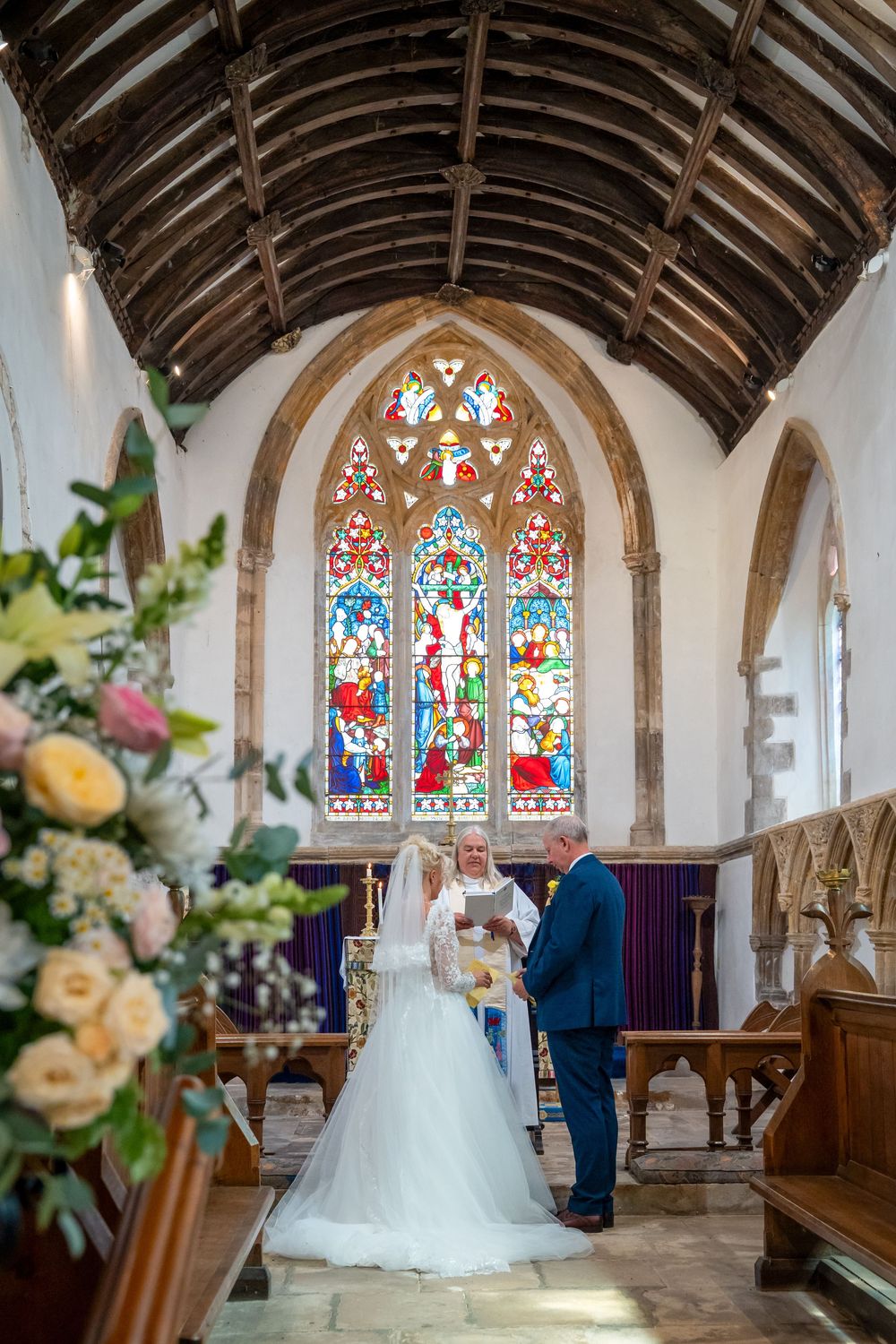 Bride and groom exchange vows at church altar with stunning stained glass windows and wooden vaulted ceiling.