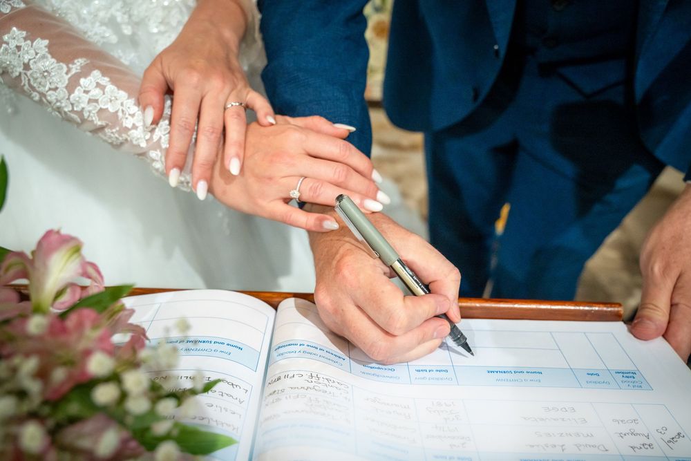 Newlyweds signing wedding registry book, bride in lace dress with rings visible, groom in blue suit holding pen.