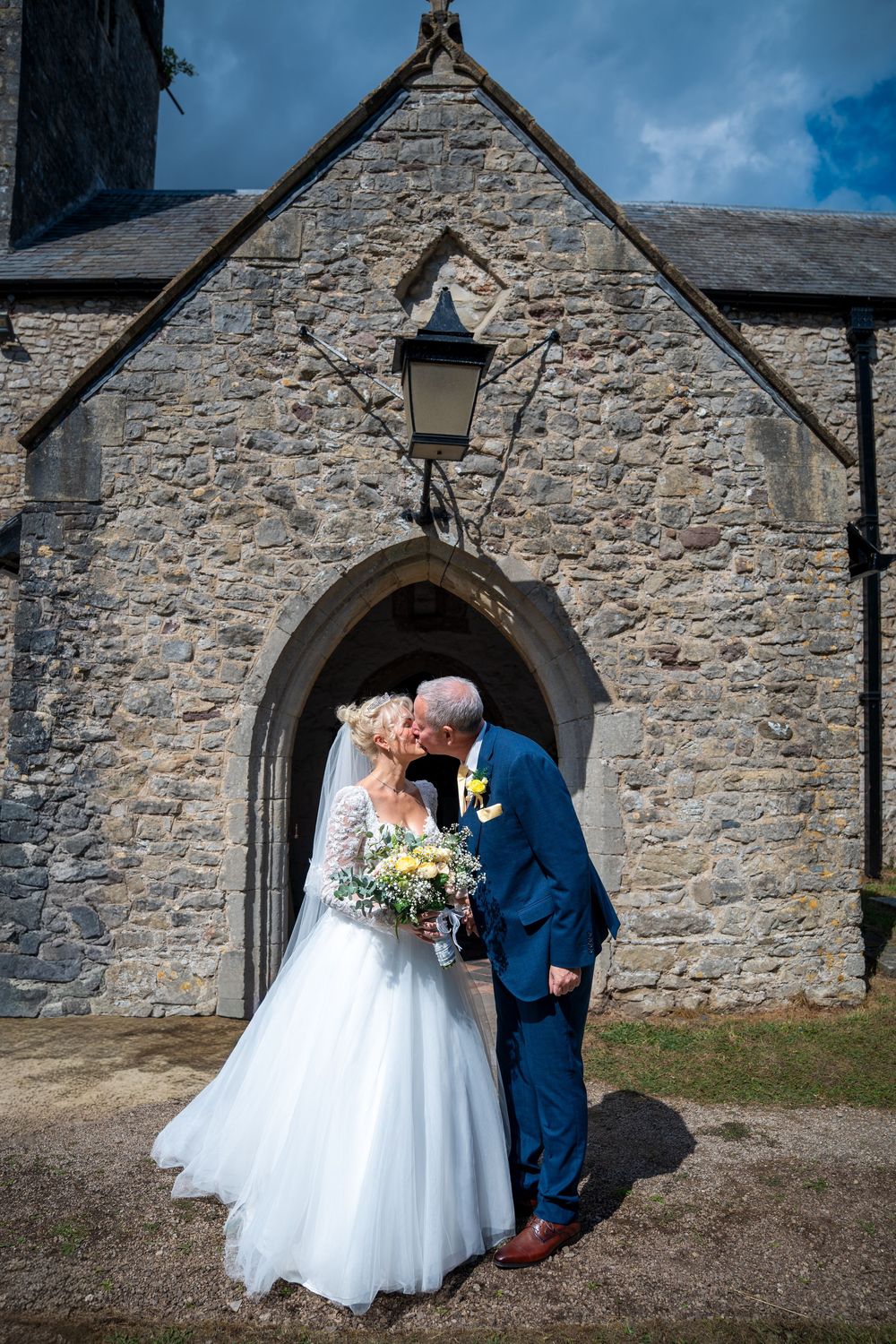 Bride and groom kissing outside a historic stone church, bride holding yellow and white bouquet in white gown.