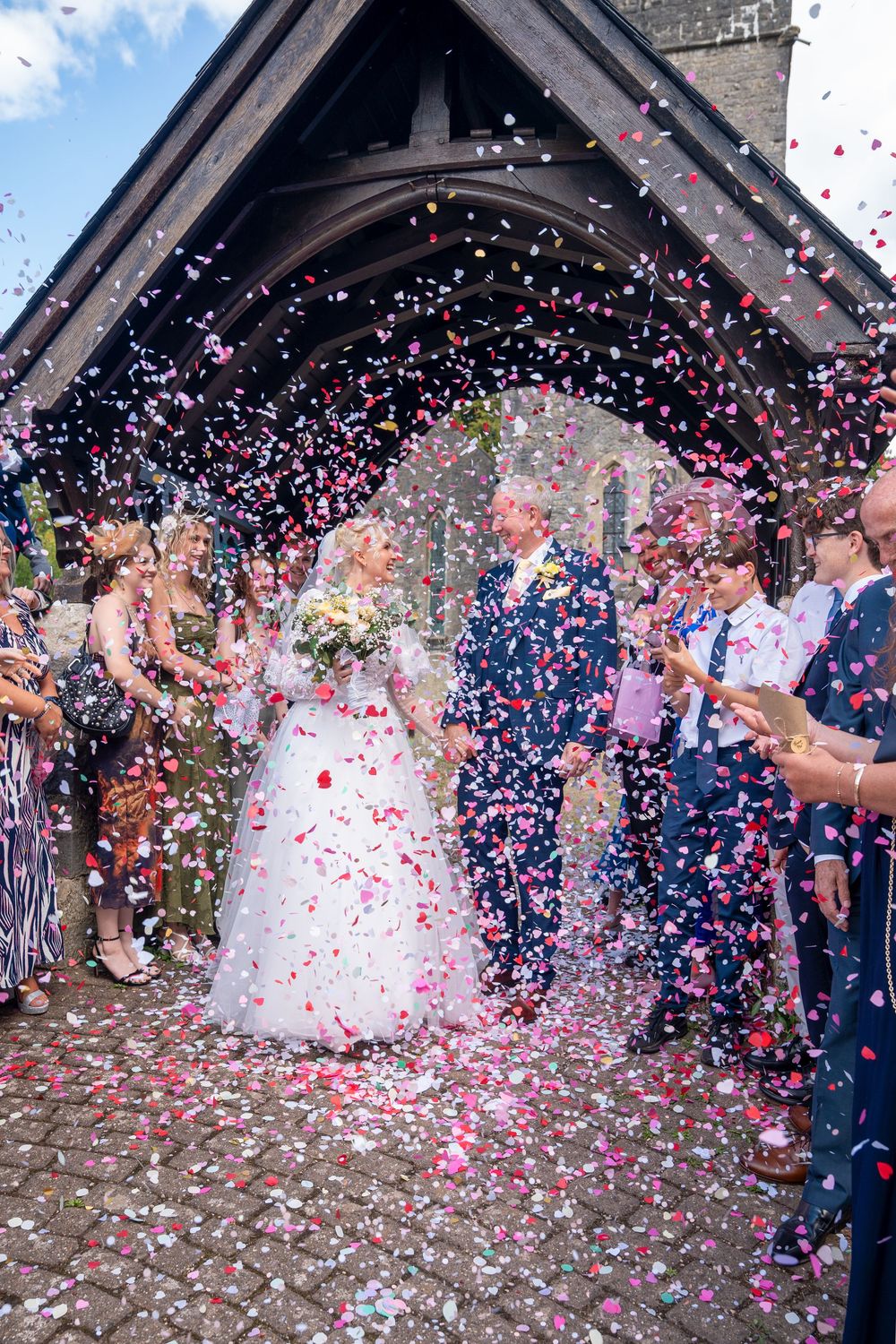 Bride and groom exit church amid colorful confetti shower, surrounded by cheering guests on their wedding day.