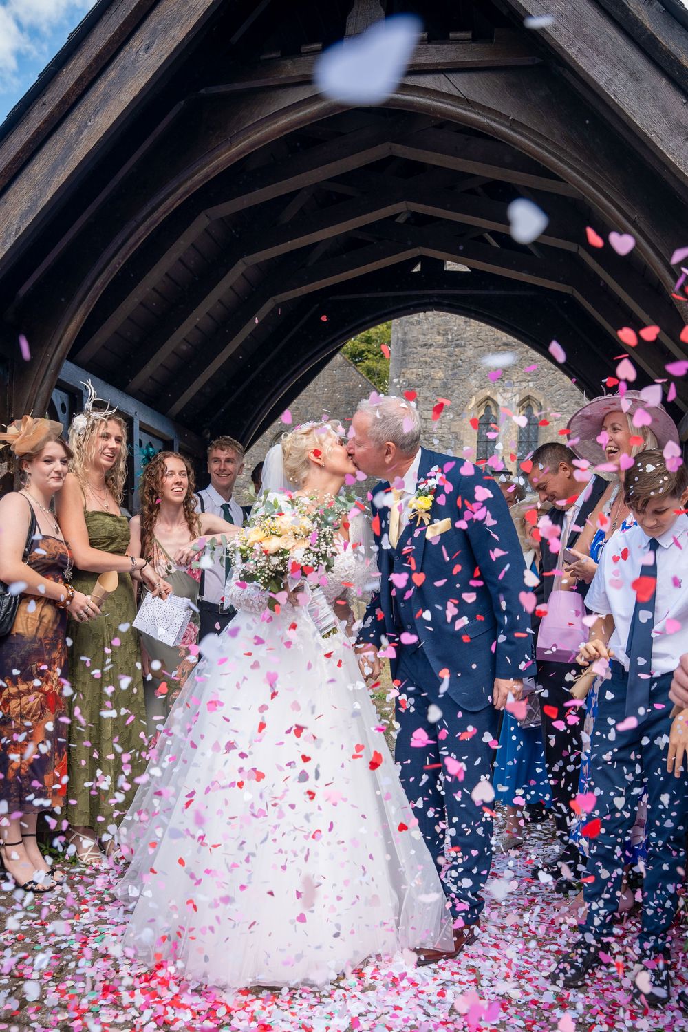 Bride and groom share a kiss under a wooden arch as guests throw colorful confetti at their wedding ceremony.