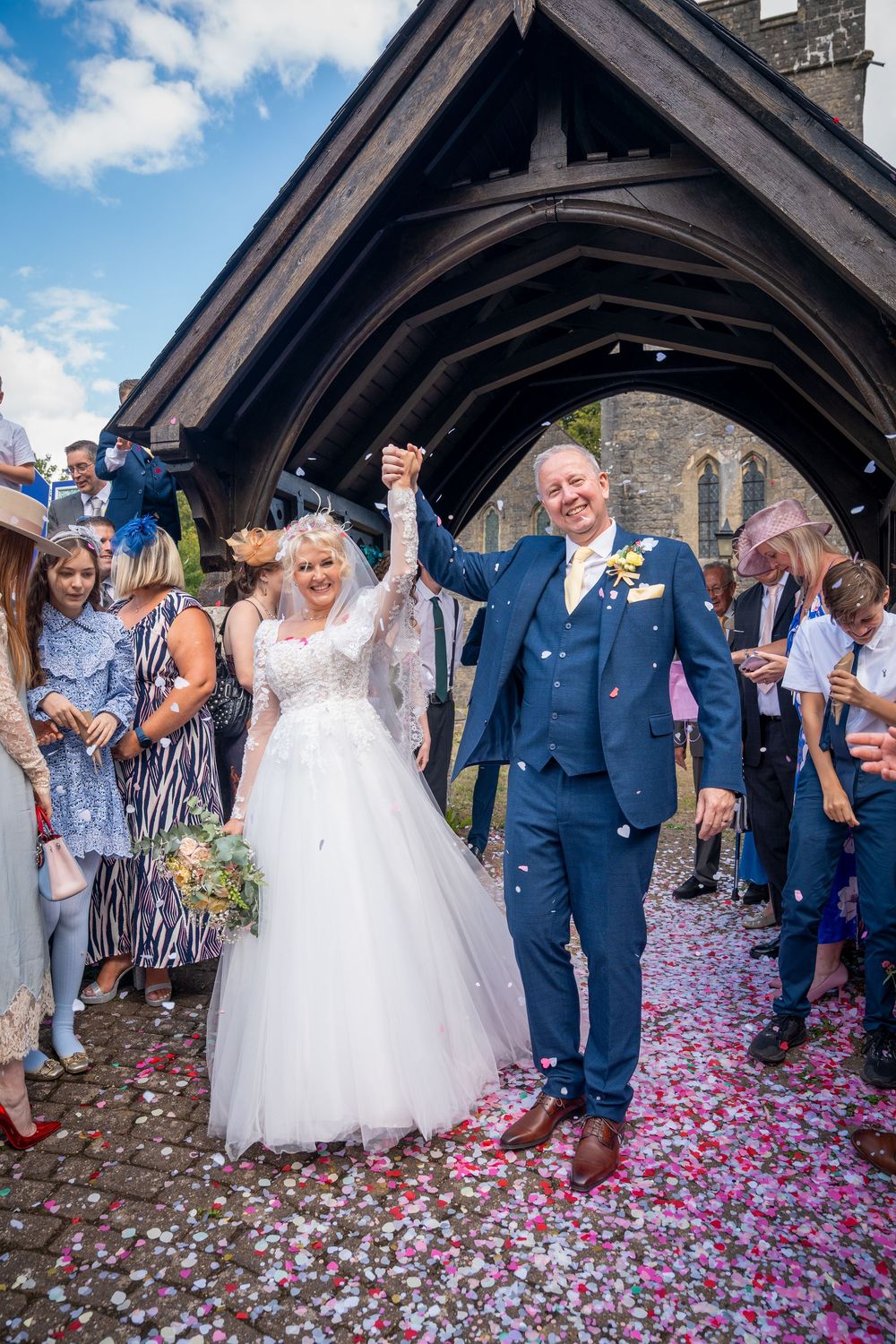 Bride and groom celebrate with raised hands as guests throw colorful confetti outside a historic church.