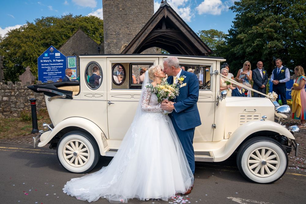 Bride and groom share a kiss beside a vintage cream wedding car outside a historic church.
