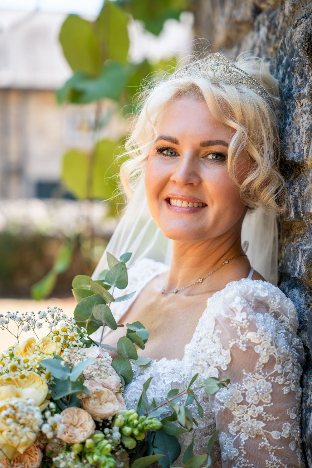 Smiling blonde bride in lace wedding dress holding bouquet of roses and eucalyptus, leaning against stone wall outdoors.