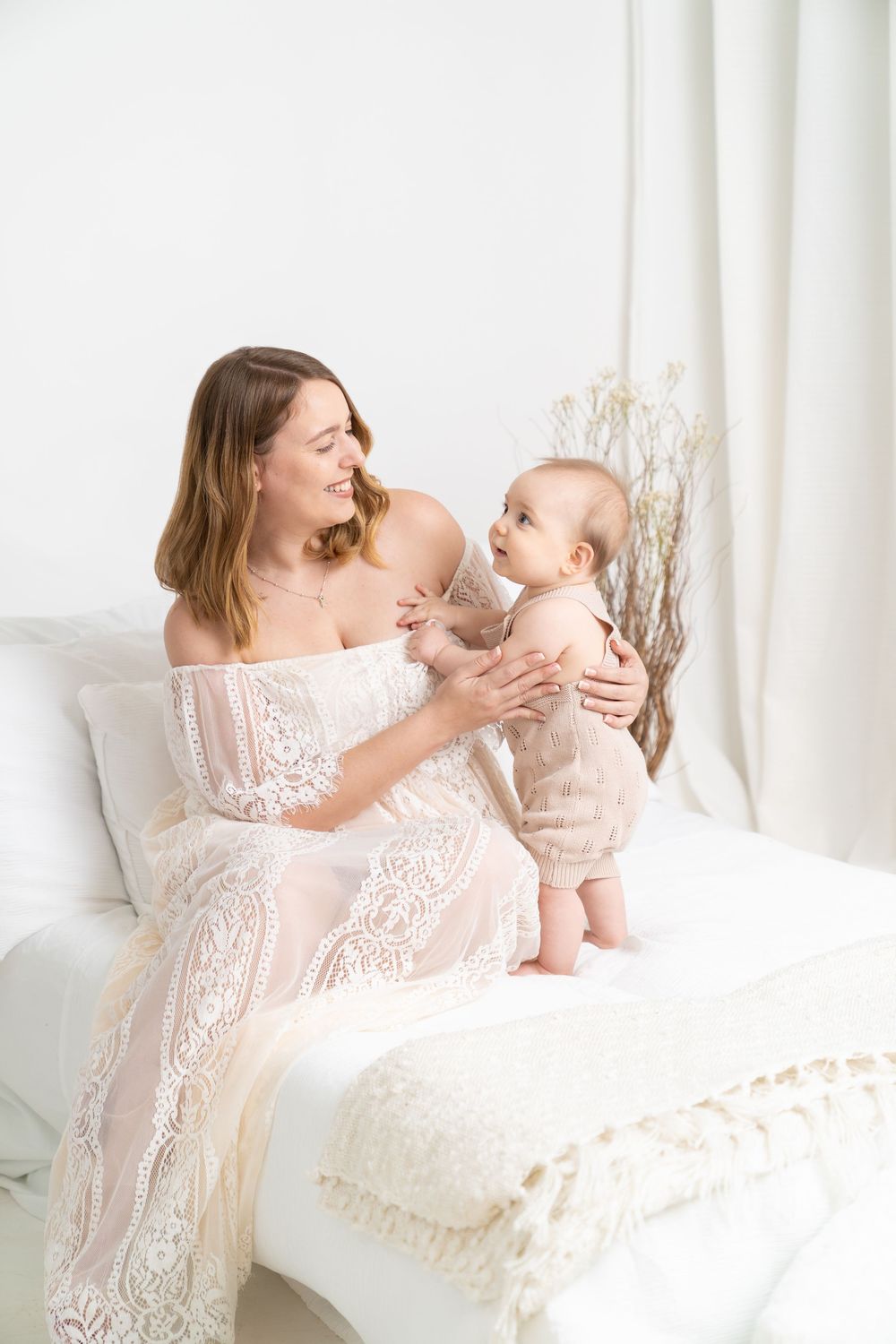 A smiling mother in a white lace dress sits on a bed holding her baby in a bright, airy white bedroom.