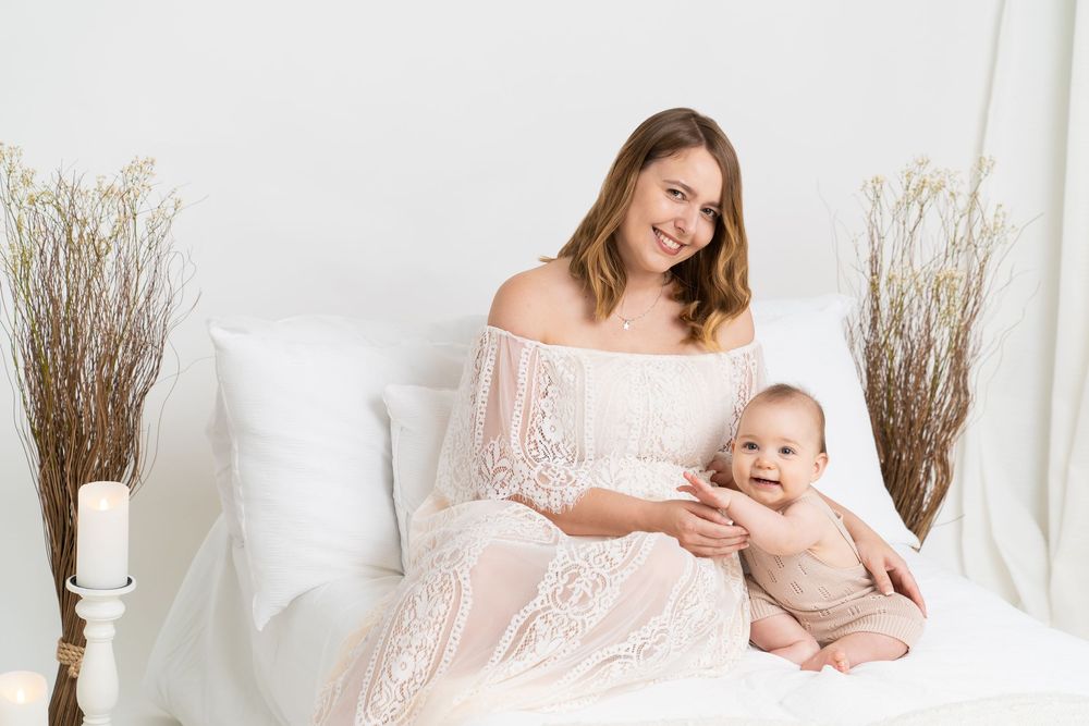 A smiling mother in a white lace dress sits on a bed holding her happy baby in a bright, elegant white room.