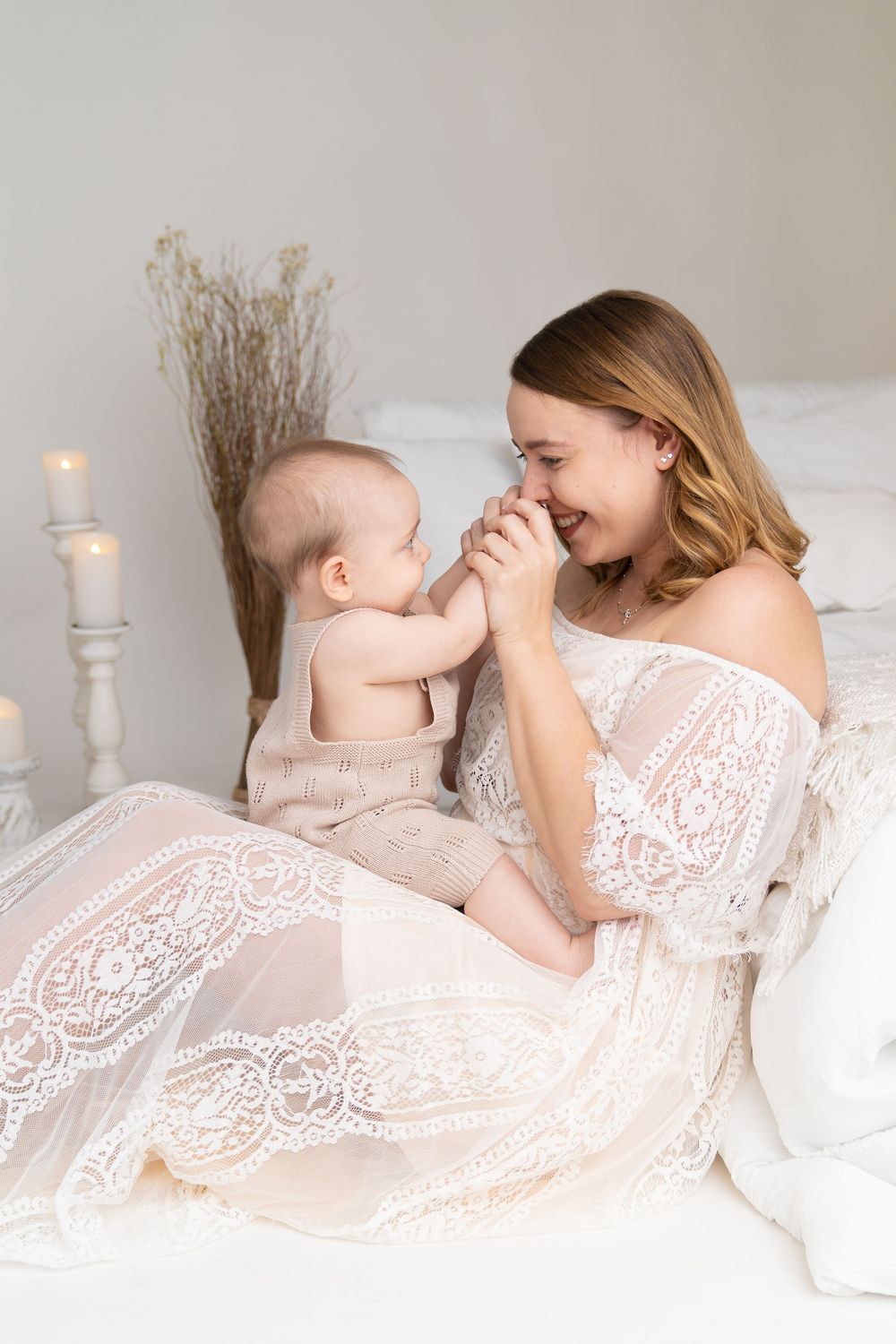 A young mother in a white lace dress sits on a bed, lovingly interacting with her baby in a soft, candlelit room.