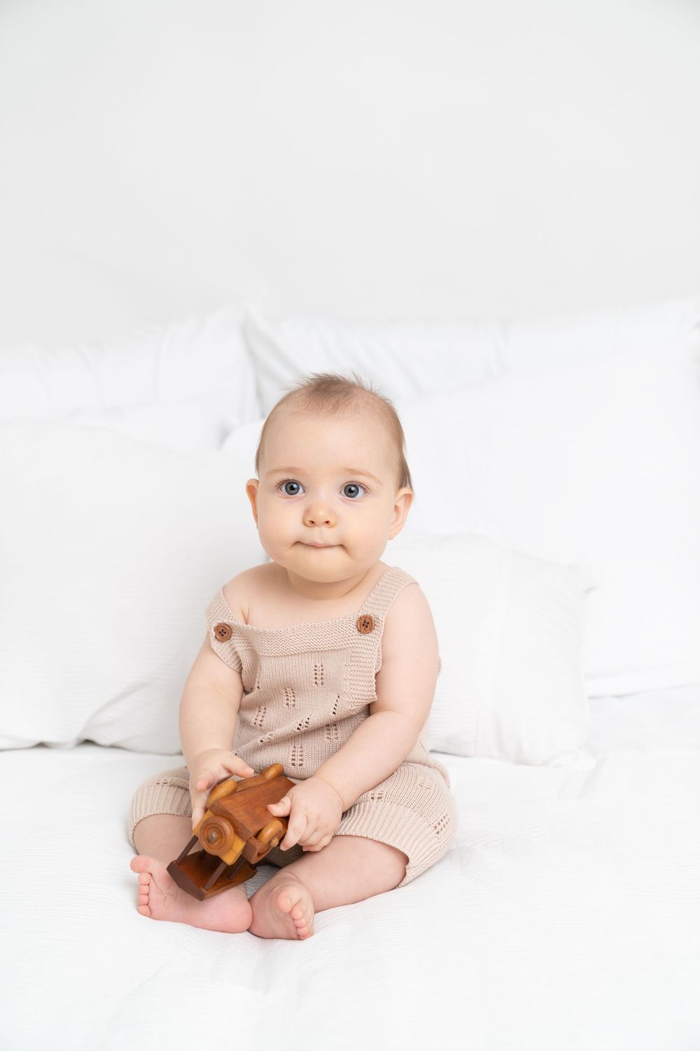 Cute baby in beige knit romper sitting on white bed holding a wooden toy, looking at camera.