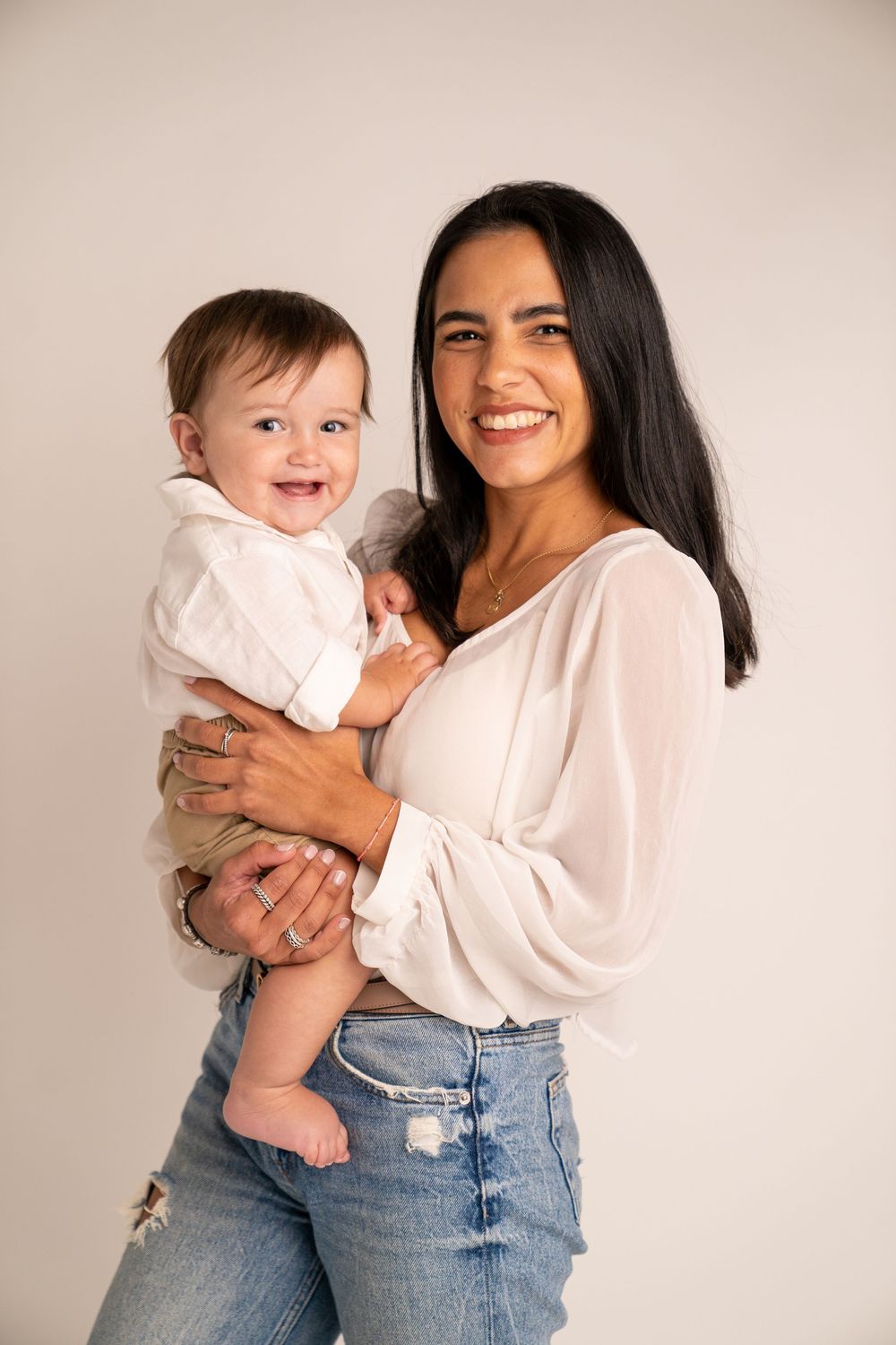 Smiling woman in white blouse and jeans holding a happy baby in a white outfit against a neutral background.