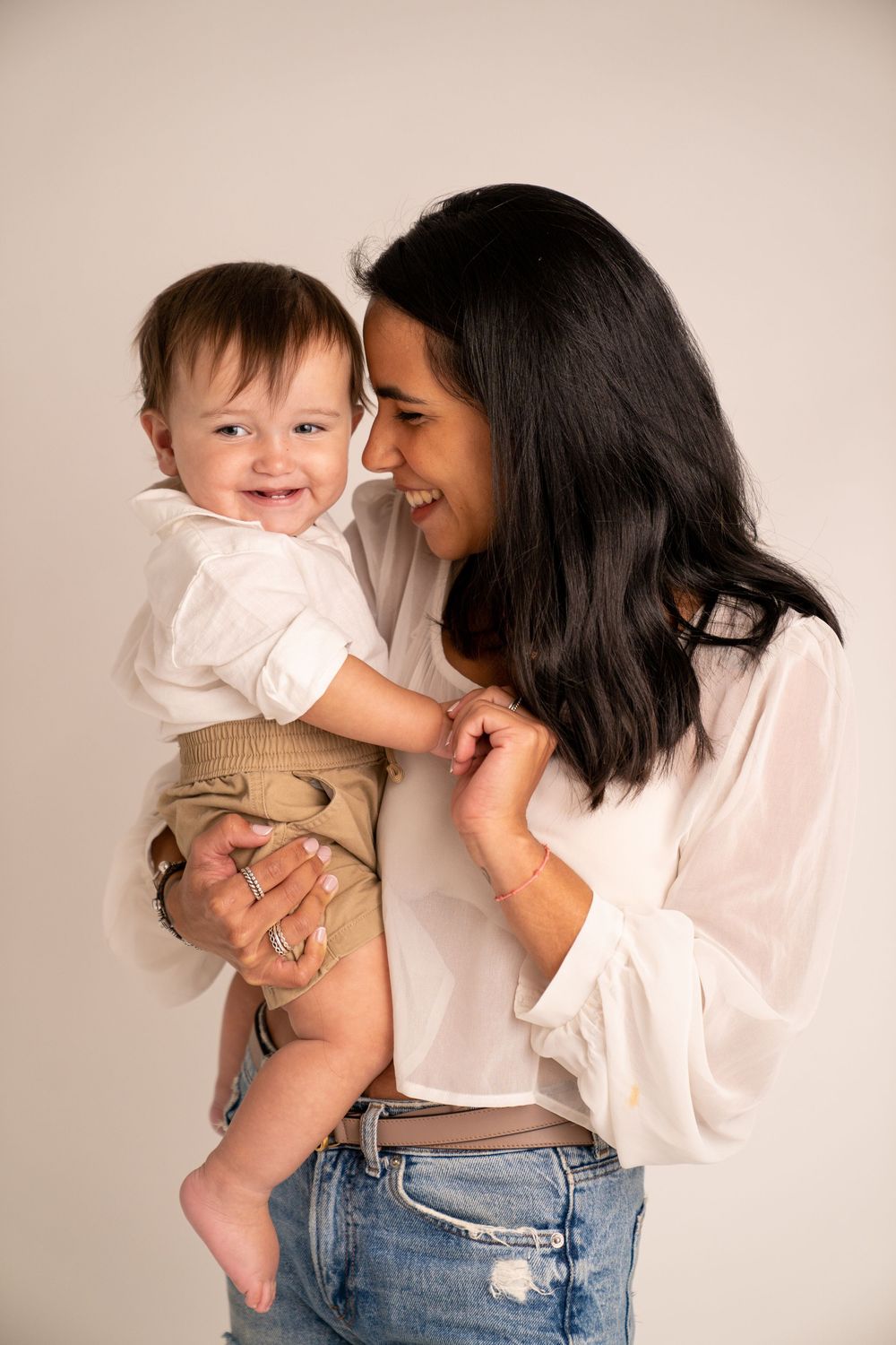 Smiling dark-haired mother in white blouse holds laughing baby boy in tan shorts against neutral backdrop.